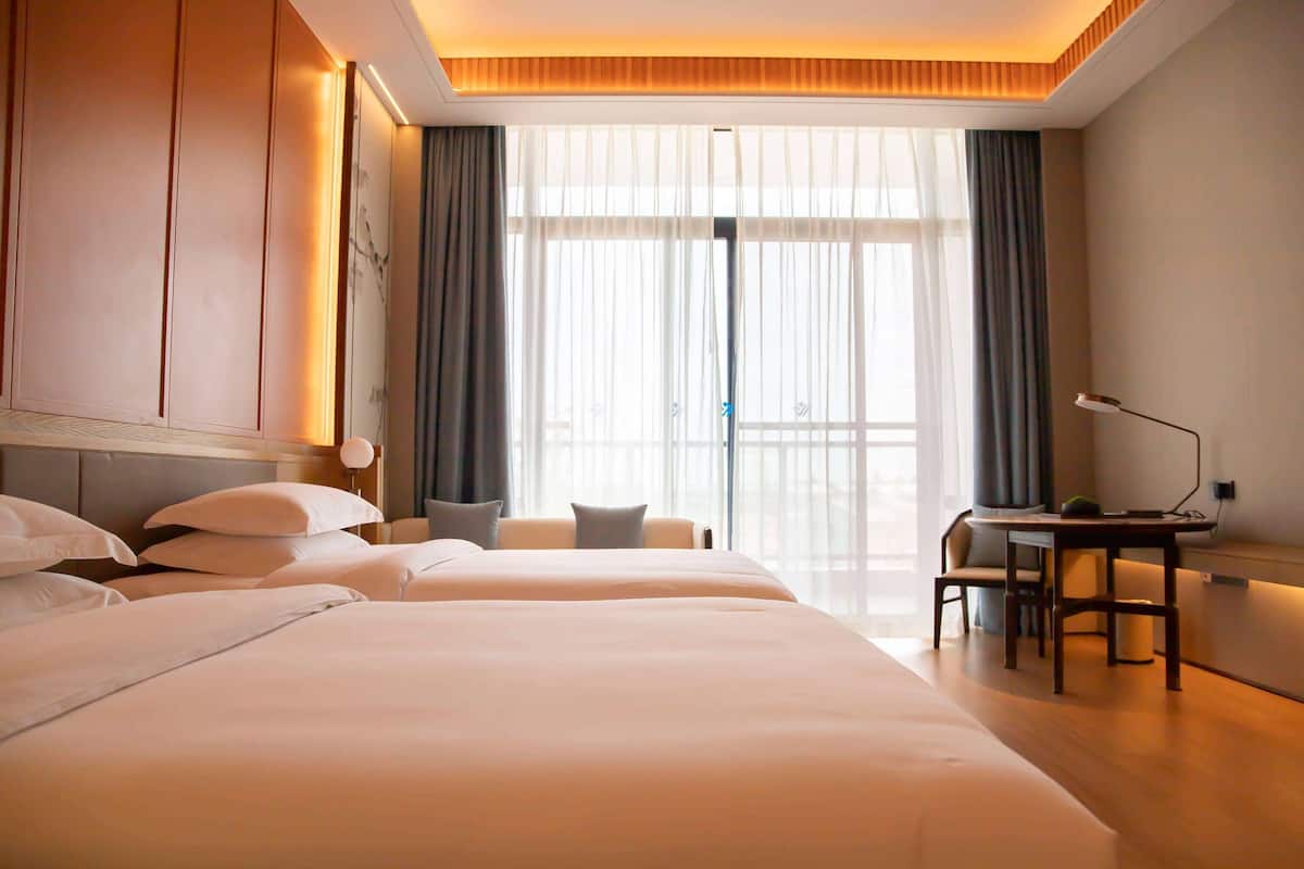 Ein Hotelzimmer mit einem großen Bett, einem Schreibtisch und einem Stuhl. Ein Fenster mit Vorhängen und Blick ins Freie.