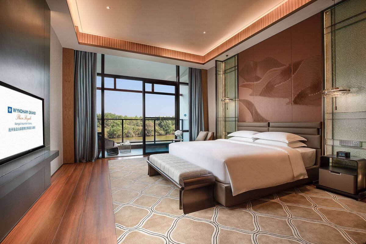 Ein modernes Hotelzimmer mit einem großen Bett, einer Bank, einem Fernseher und Blick ins Freie.
