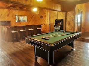 A pool table with cues and balls, wooden walls, a bar area with stools, and a door with a mirror.