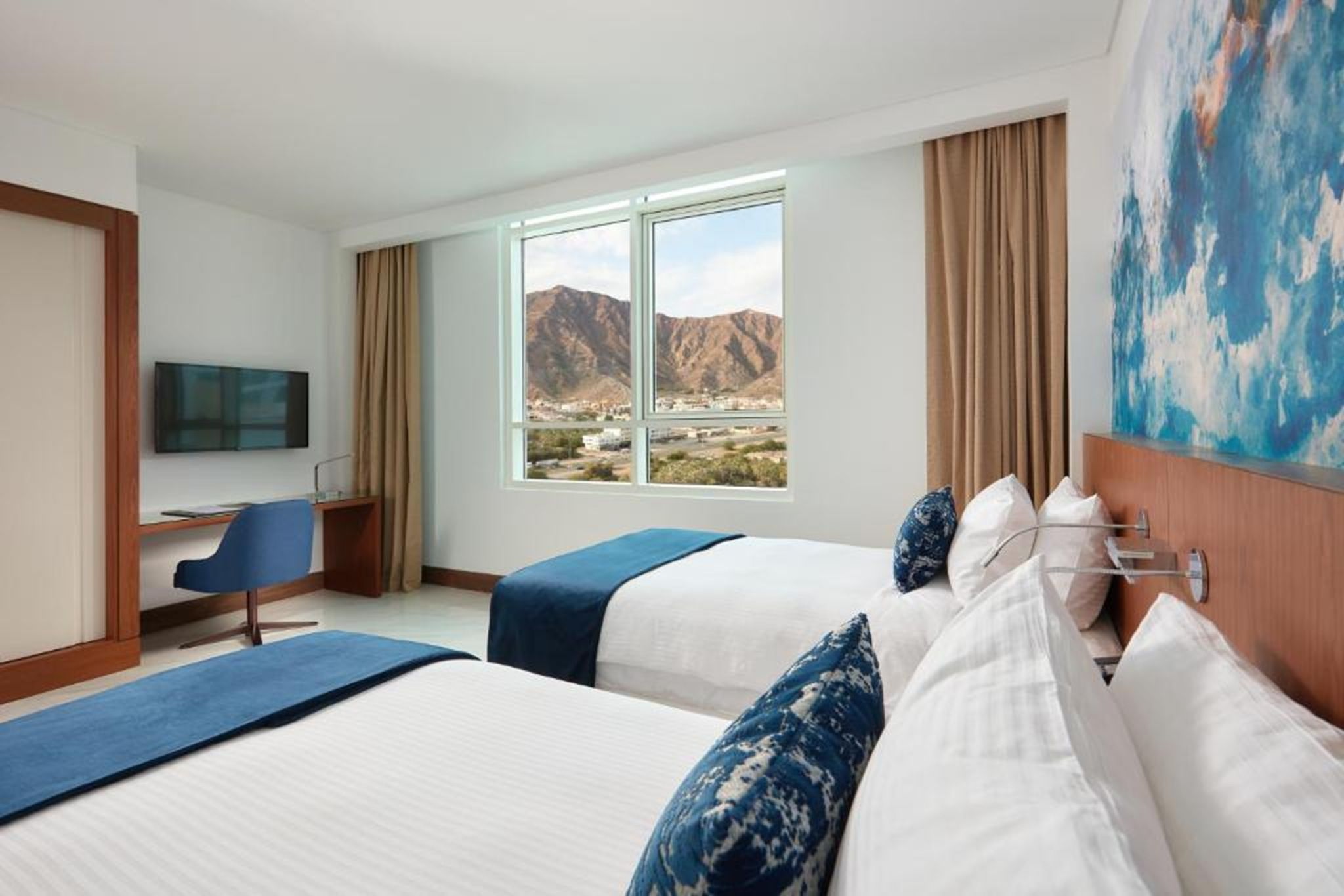 A hotel room with two beds, a desk, a chair, a TV, and a large window with a mountain view.