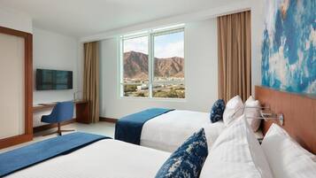 Habitación de hotel con dos camas, un escritorio, una silla, un televisor y un amplio ventanal con vistas a la montaña.