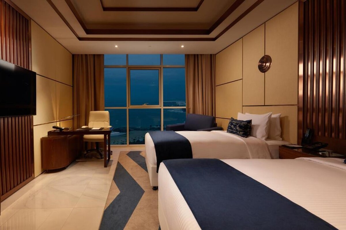A hotel room with two beds, a desk, a chair, a TV, and a large window with a view.