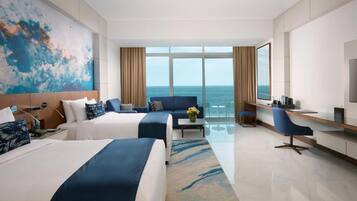 A hotel room with two beds, a desk, a chair, a TV, and a large window with a view of the sea.
