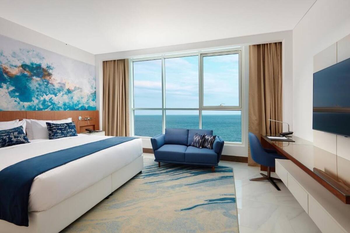 Quarto de hotel com cama, sofá, escrivaninha, televisão e uma grande janela vista para o mar.