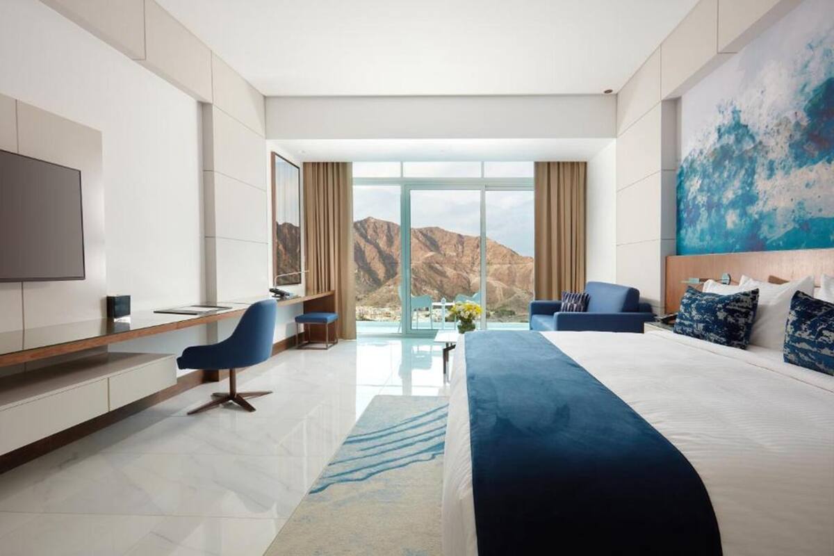 Quarto de hotel moderno com uma cama grande, uma escrivaninha, uma cadeira, uma televisão e vista para montanhas.