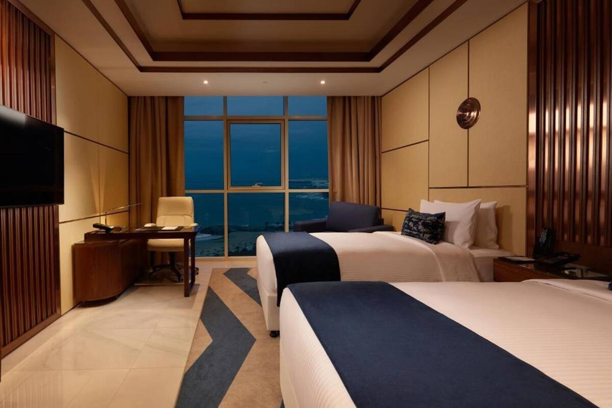 Quarto de hotel com duas camas, uma escrivaninha, uma cadeira, televisão e uma janela grande com vista.