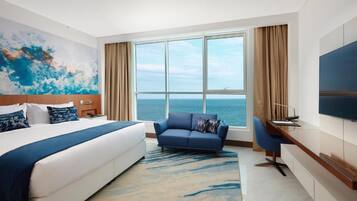 A hotel room with a bed, a sofa, a desk, a TV, and a large window with a view of the sea.