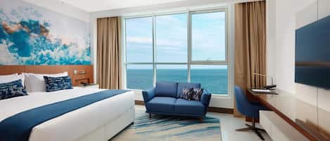 A hotel room with a bed, a sofa, a desk, a TV, and a large window with a view of the sea.