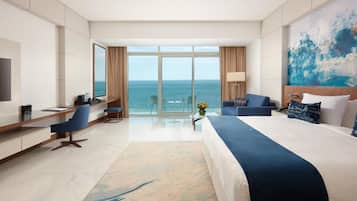 A hotel room with a large bed, a desk, a chair, a TV, and a view of the ocean.