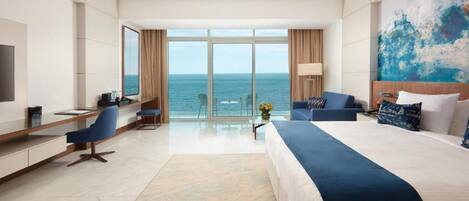 A hotel room with a large bed, a desk, a chair, a TV, and a view of the ocean.