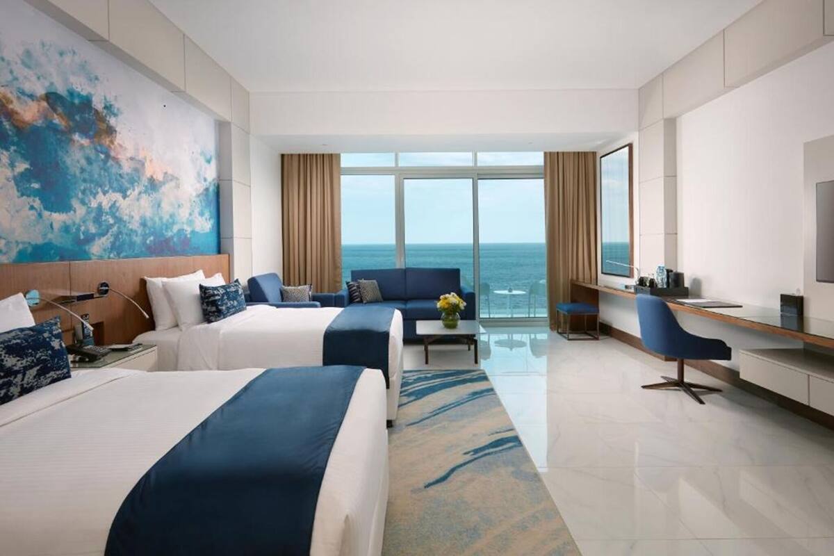 Quarto de hotel com duas camas, uma escrivaninha, uma cadeira, televisão e uma janela grande com vista para o mar.