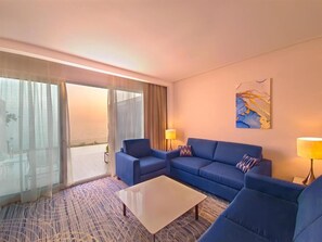 A hotel room with a large sliding glass door to a balcony, a blue sofa set, a small coffee table, and a painting on the wall. - Royal M Al Aqah Beach Hotel And Resort by Gewan (Al Aqah)