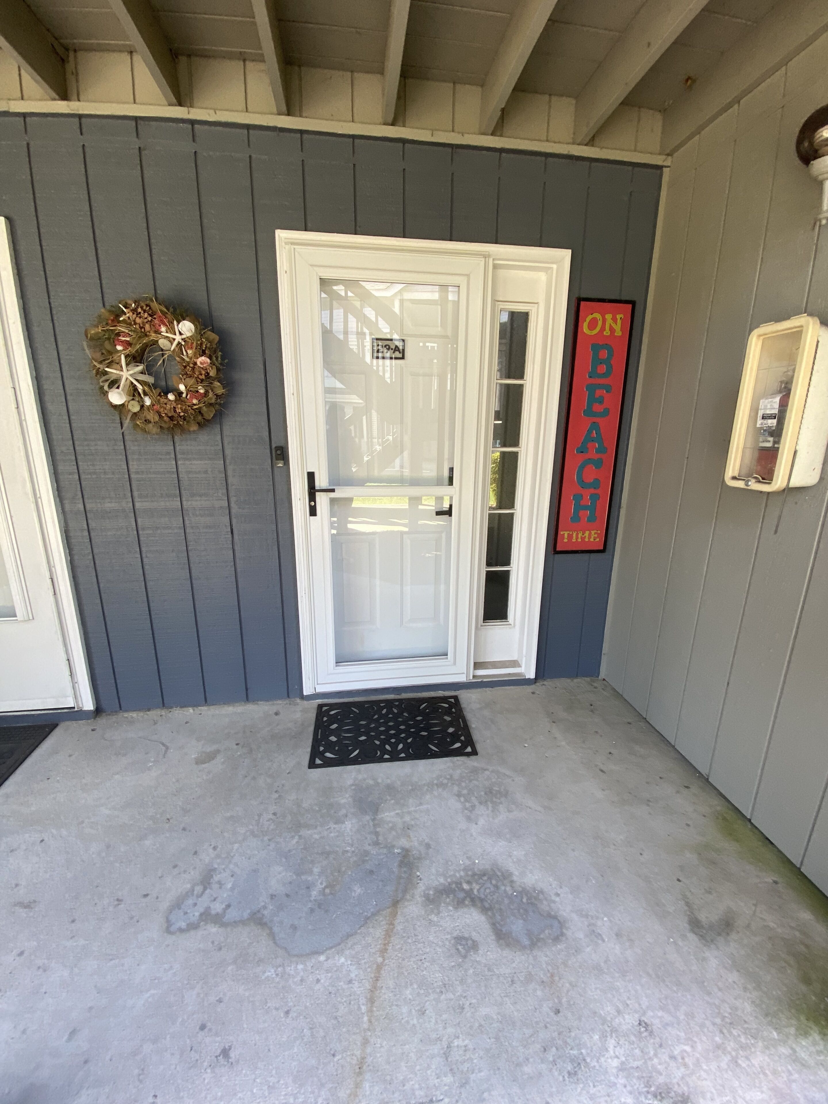 A front door with a glass panel, a wreath, a 'ON BEACH TIME' sign, and a doormat.