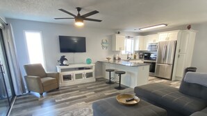 A modern living room with a grey sofa, a coffee table, and a TV mounted on the wall. There is a kitchen area with white cabinets and stainless steel appliances. - Minutes to beach and golf courses (Myrtle BEach)