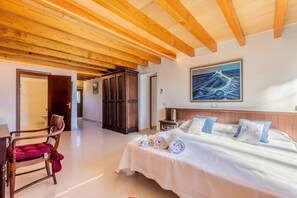 A bedroom with a wooden ceiling, a bed with white bedding and blue pillows, a nightstand, and a painting of waves on the wall.
