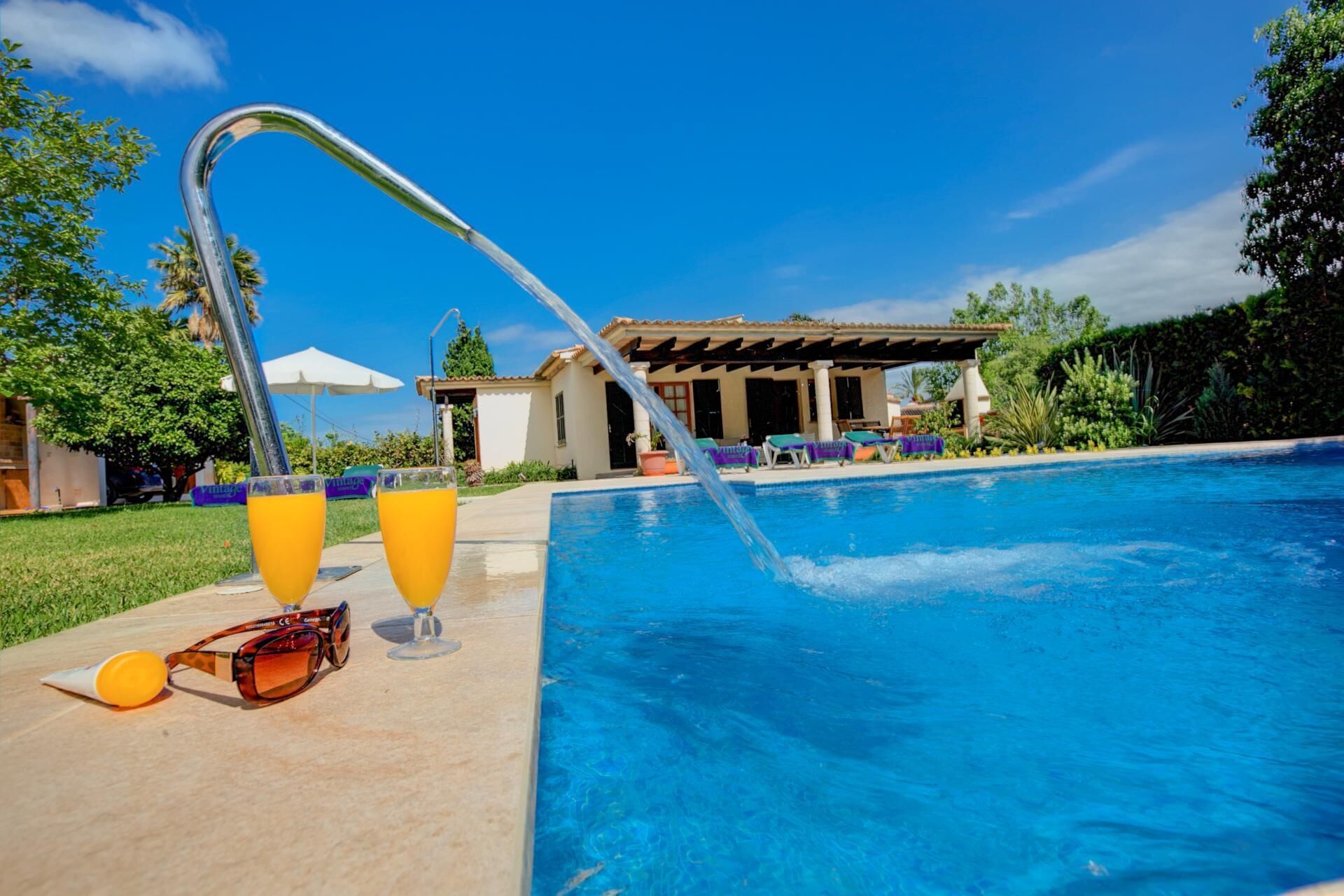 Ein Schwimmbecken mit einer Wassersäule, zwei Orangensaftgläsern und einer Sonnenbrille auf der Poolterrasse.