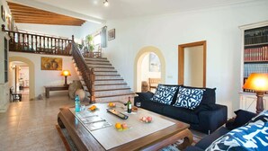 A spacious living room with a wooden staircase, a black sofa with patterned cushions, a coffee table with magazines and fruit, and a bookshelf filled with books. - Fantastic Spacious Villa, A/C, Big Private Pool, Idyllic Location - 5/10 mins to Towns & Beaches! (Alcudia)