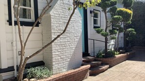 A white building with a brick base, a window with black shutters, and a small tree in a planter. - The Boardwalk Beach House (Santa Cruz)