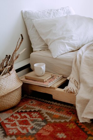 A neatly made bed with a white pillow and a white blanket, a white mug, a book, and a woven basket on a wooden nightstand. - The Boardwalk Beach House (Santa Cruz)