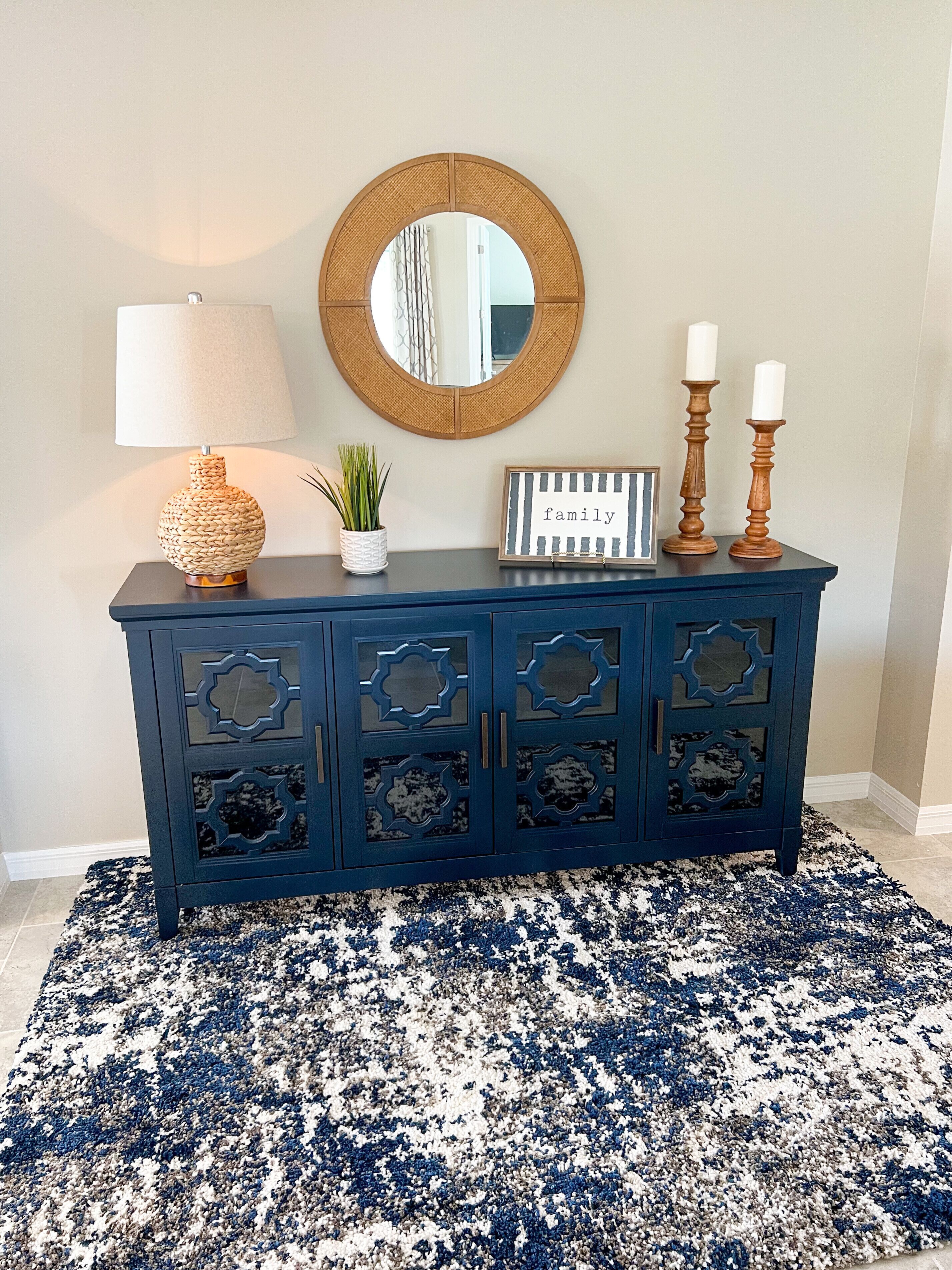 Un mueble azul oscuro con adornos recortados, un espejo redondo, una lámpara blanca, una pequeña planta en maceta y dos candelabros con velas blancas.