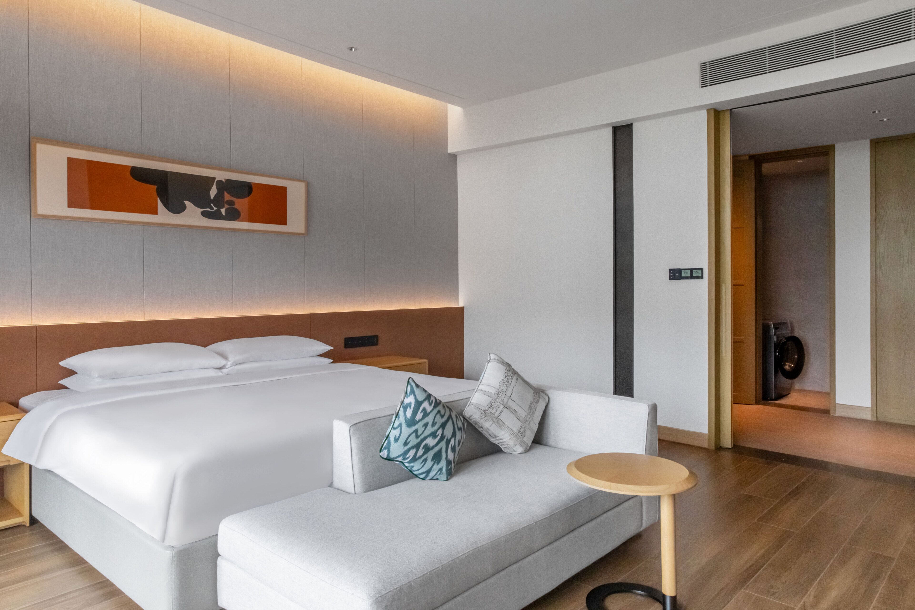 A modern hotel room with a large bed, a sofa, a small round table, and a wall-mounted artwork.
