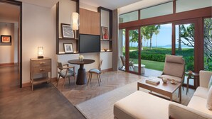 Un saló modern amb una finestra gran que ofereix vistes a un paisatge tropical. Hi ha un sofà, una taula de menjador amb cadires, i una taula auxiliar petita amb un llibre i una tetera.