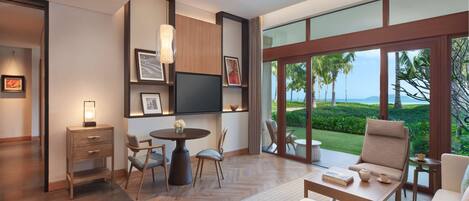 A modern living room with a large window offering a view of a tropical landscape. There is a sofa, a dining table with chairs, and a small side table with a book and a teapot.