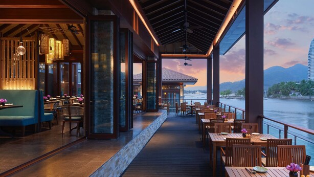 Un restaurant al costat del mar amb taules i cadires de fusta, un passadís cobert i grans finestres que ofereixen vistes a l'aigua i al perfil urbà.