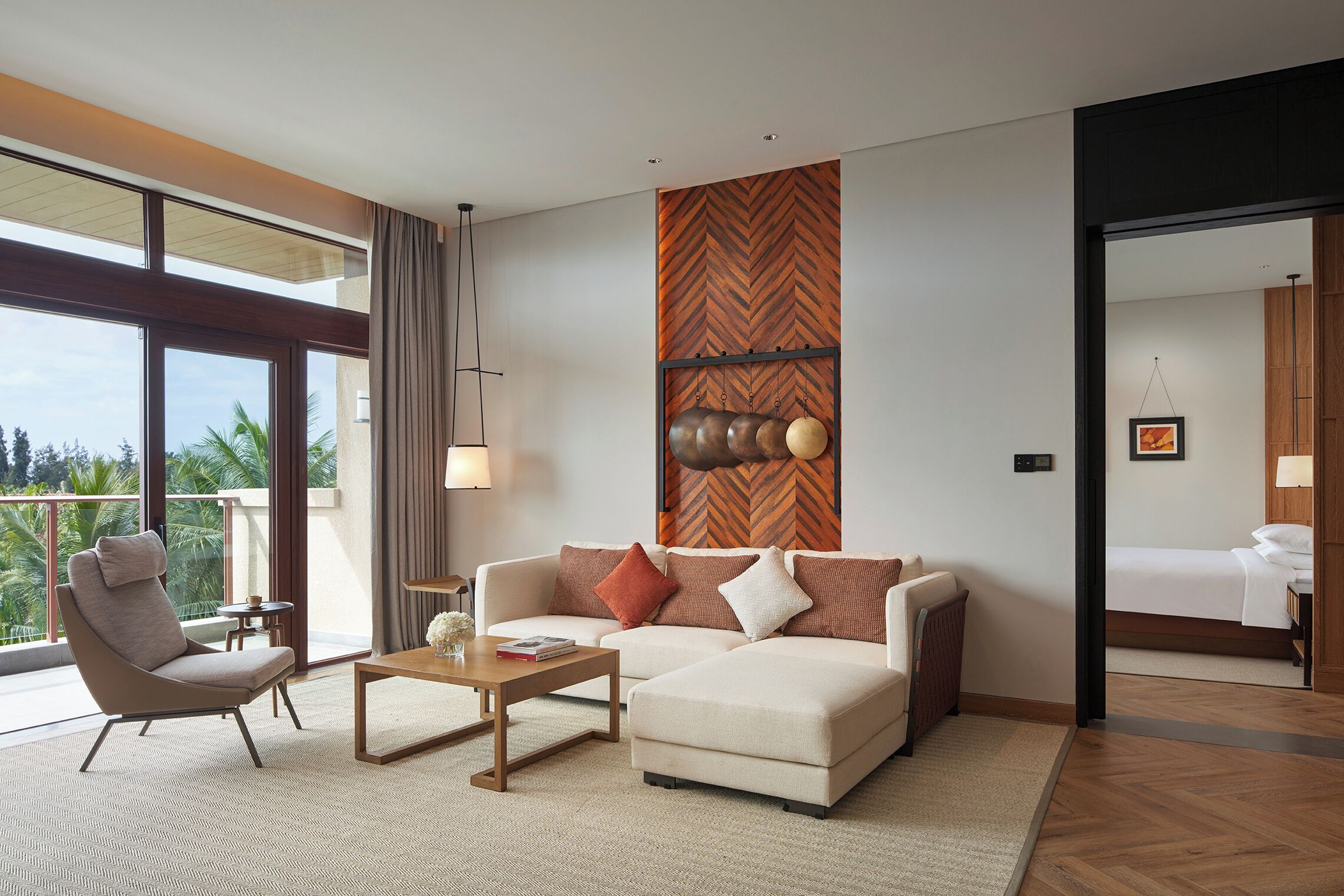 A modern hotel room with a sofa, armchair, and a small table. There is a large window offering a view of palm trees.