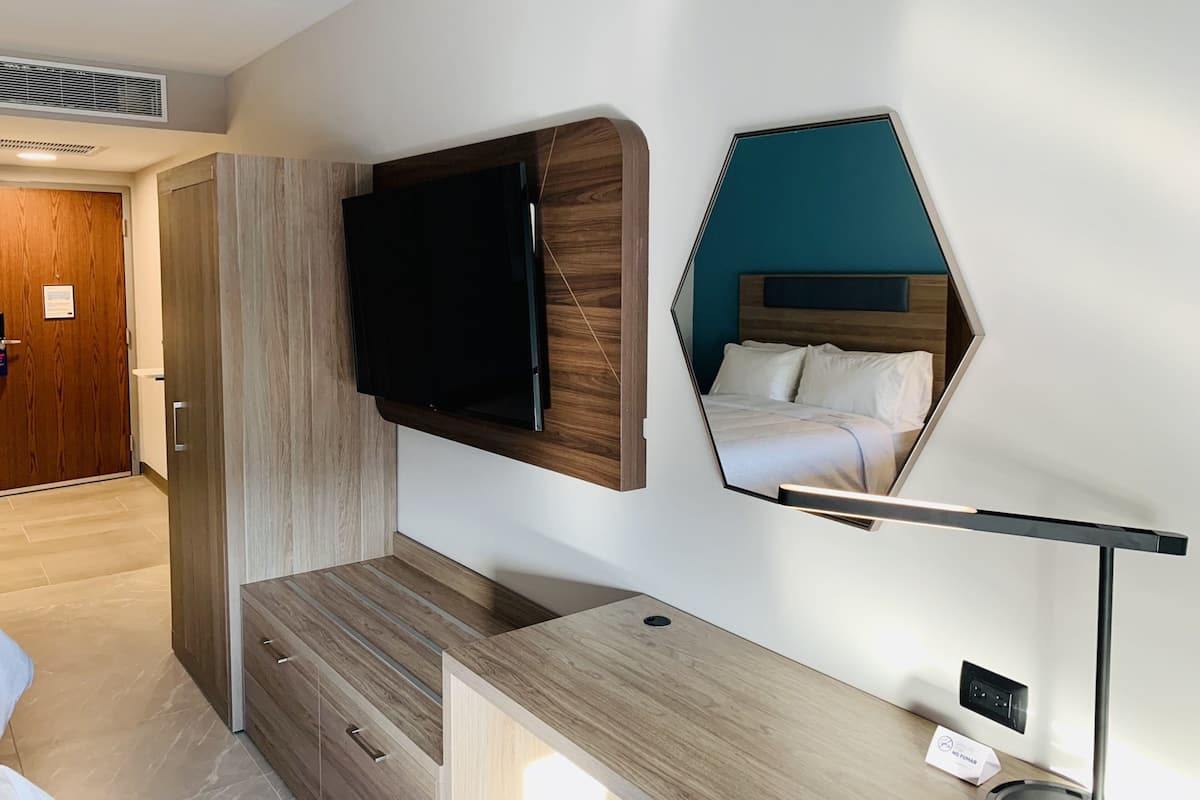 A modern hotel room with a flat-screen TV mounted on a wooden panel, a mirrored headboard reflecting the bed, and a bedside table with a lamp.