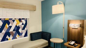 Una habitación de hotel moderna con una pared de diseño geométrico, un sofá color beige, una pequeña mesa redonda, una mesita de noche con una lámpara y una puerta de madera con un panel luminoso.