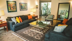Un salon avec un canapé, des fauteuils, une table basse et un tapis à motifs. Deux tableaux ornent le mur, une lampe est posée sur une table d’appoint, et une grande fenêtre offre une vue sur la verdure.