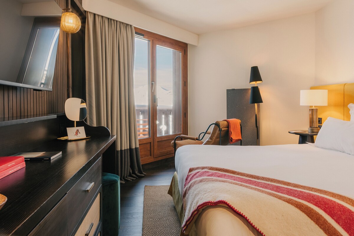 A hotel room with a bed, a desk, a chair, a lamp, a nightstand, and a view of the outdoors.