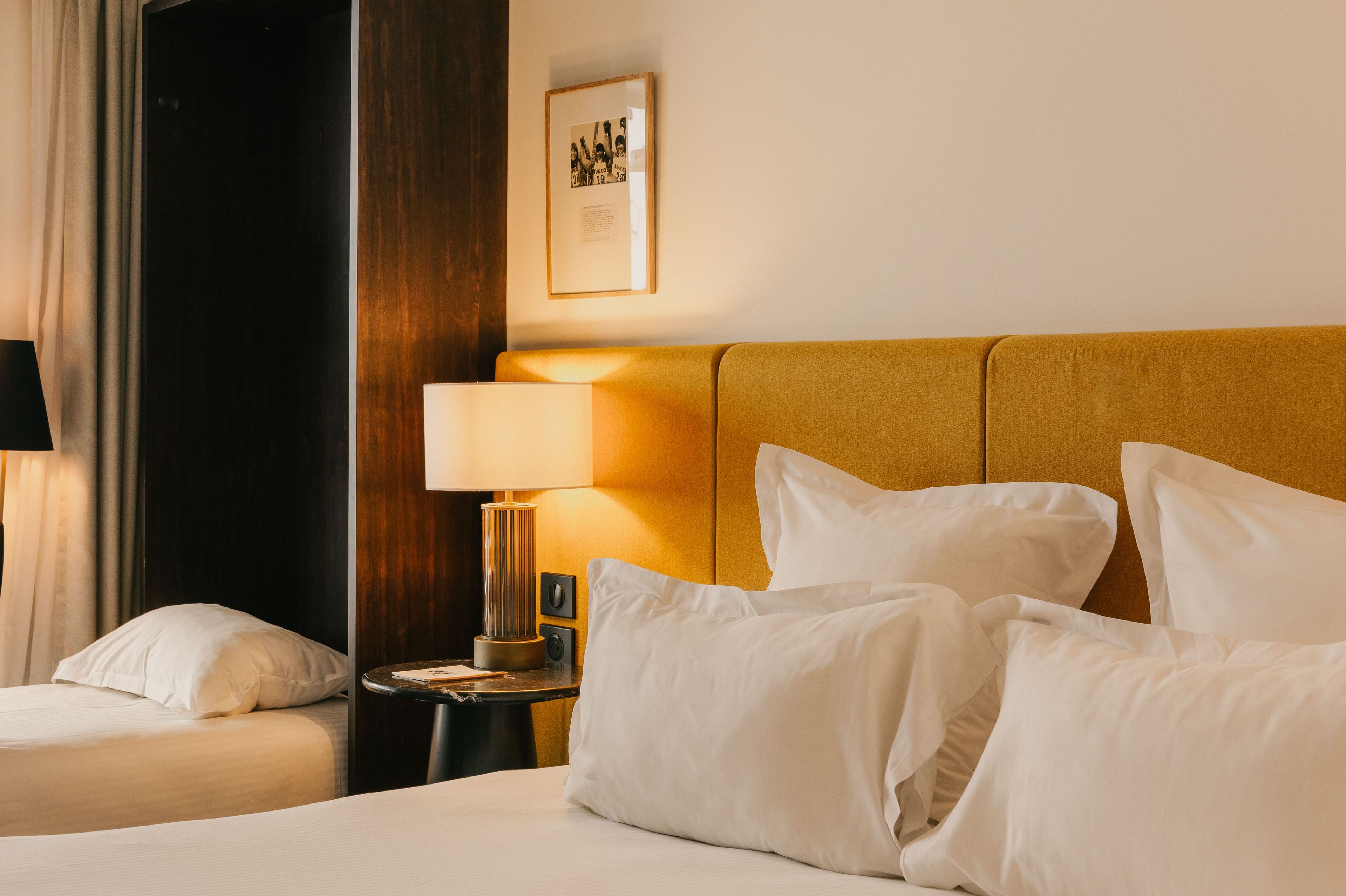 Habitación de hotel con una cama, almohadas, una mesita de noche con una lámpara, un cuadro en la pared y una puerta oscura.