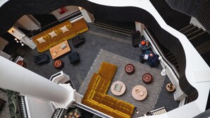 Een atrium met hoge plafonds en een centraal zitgedeelte met een grote bank en kleinere ronde tafels, omringd door een moderne trap.