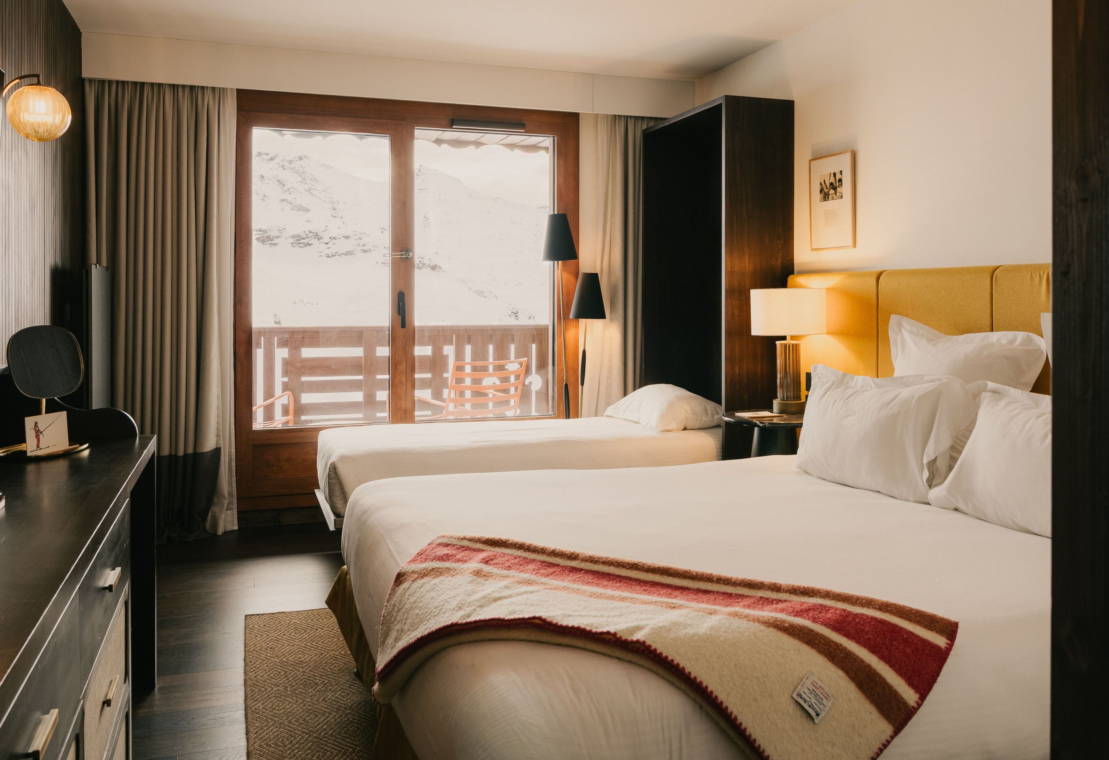A hotel room with two beds, a desk, a chair, a lamp, a nightstand, and a view of mountains through the window.