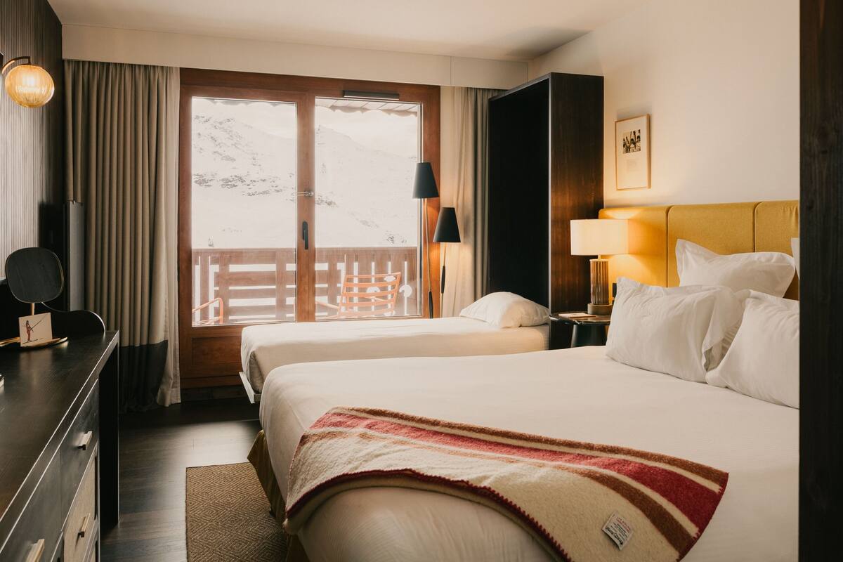 A hotel room with two beds, a desk, a chair, a lamp, a nightstand, and a view of mountains through the window.