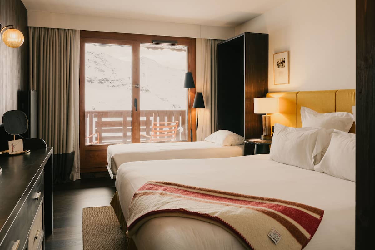 Une chambre d’hôtel avec deux lits, un bureau, une chaise, une lampe, une table de chevet et une vue sur les montagnes par la fenêtre.