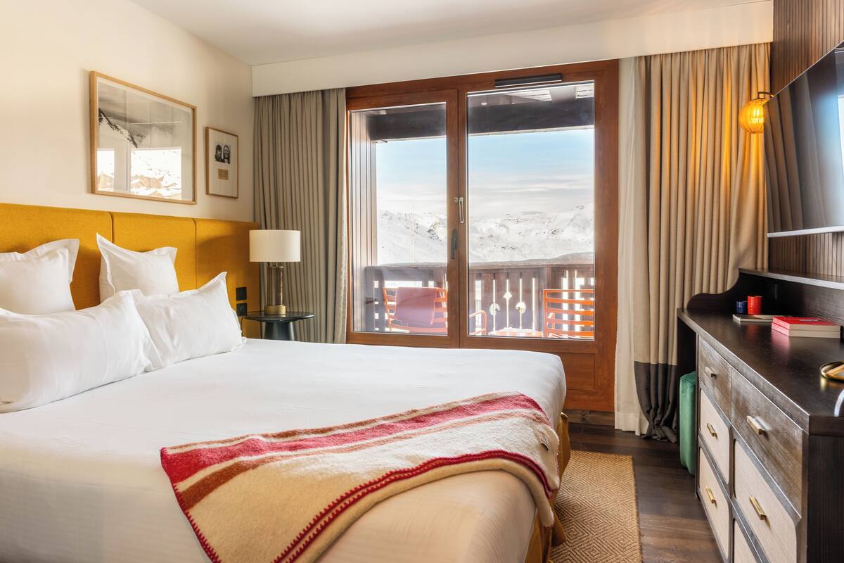 A hotel room with a large bed, a wooden headboard, a desk with a lamp, a dresser, and a view of a snowy landscape through a sliding door.