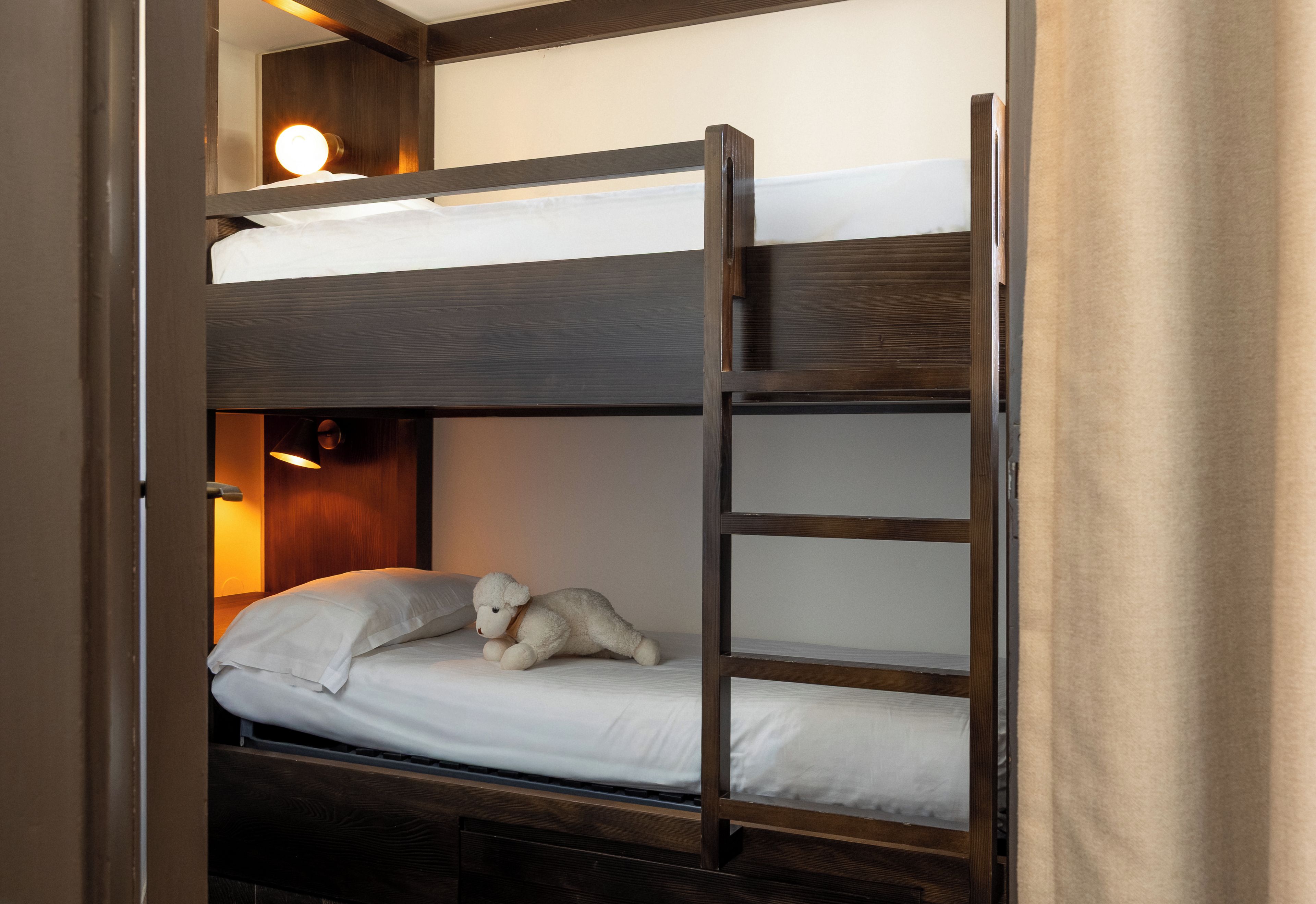 Una cama litera con un oso de peluche en la cama inferior, una lámpara fijada a la pared y un cabecero de madera.