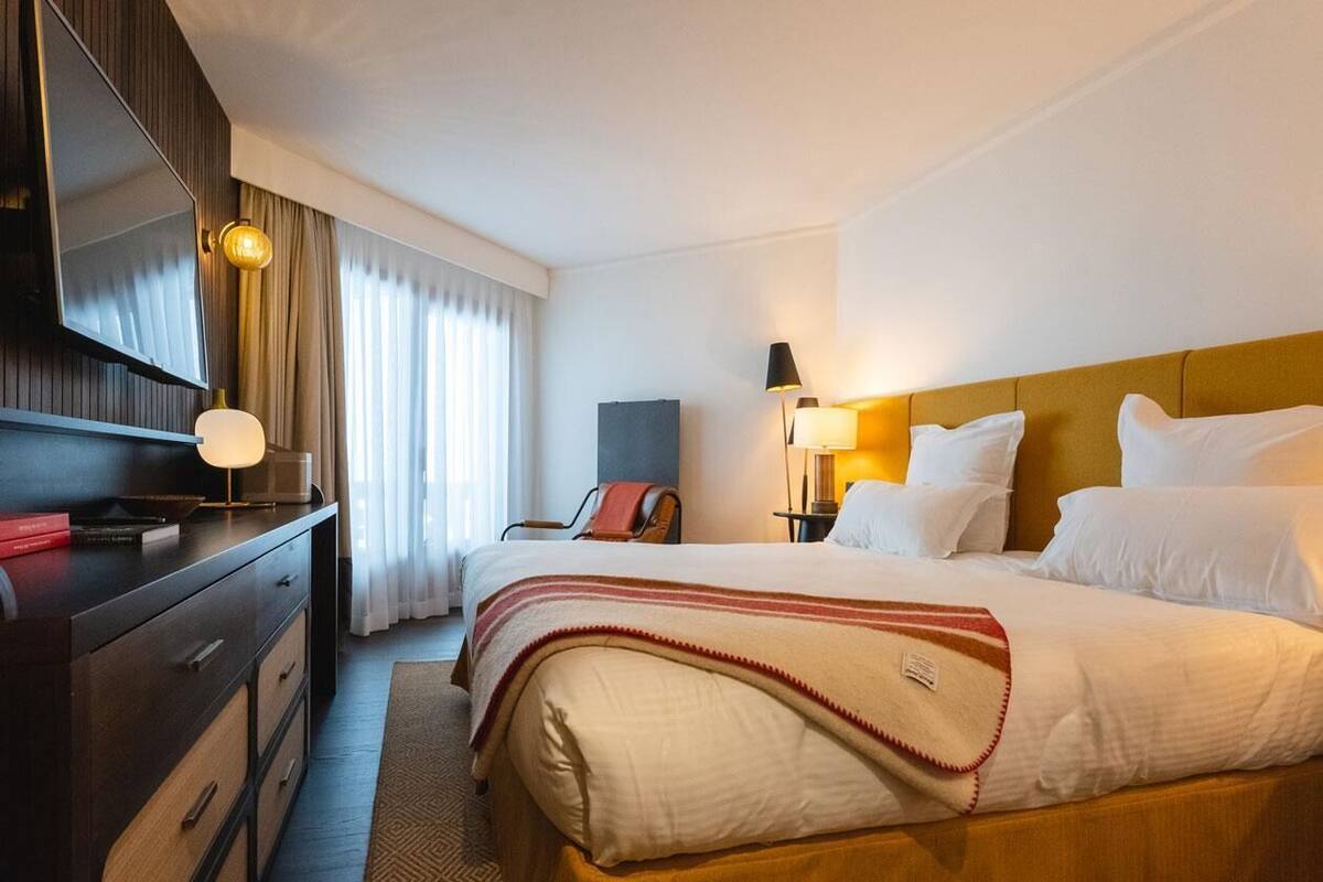 A hotel room with a bed, a desk with a lamp, a TV, and a window with curtains.