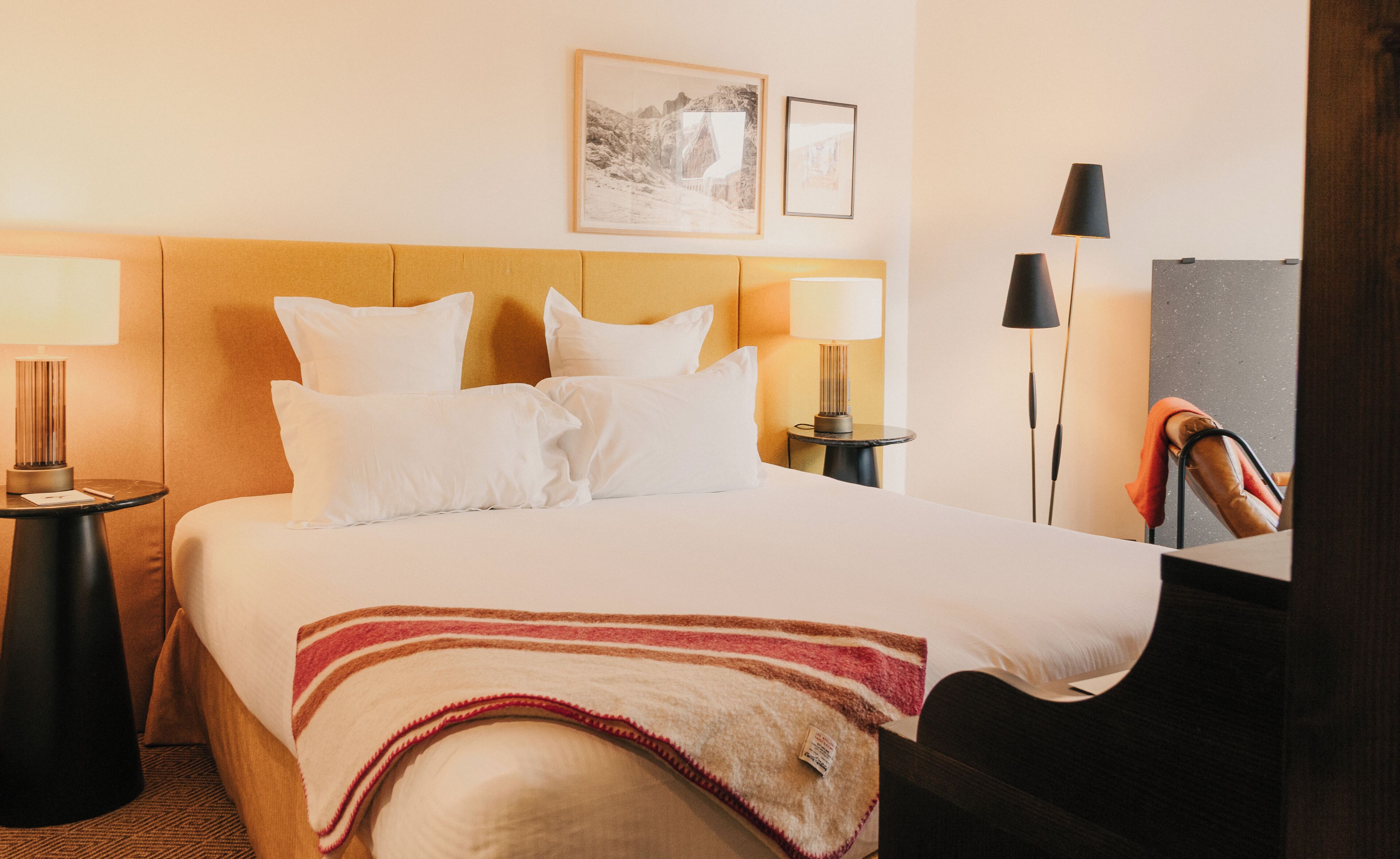 A hotel room with a bed, two bedside tables, a lamp, a chair, and two framed pictures on the wall.