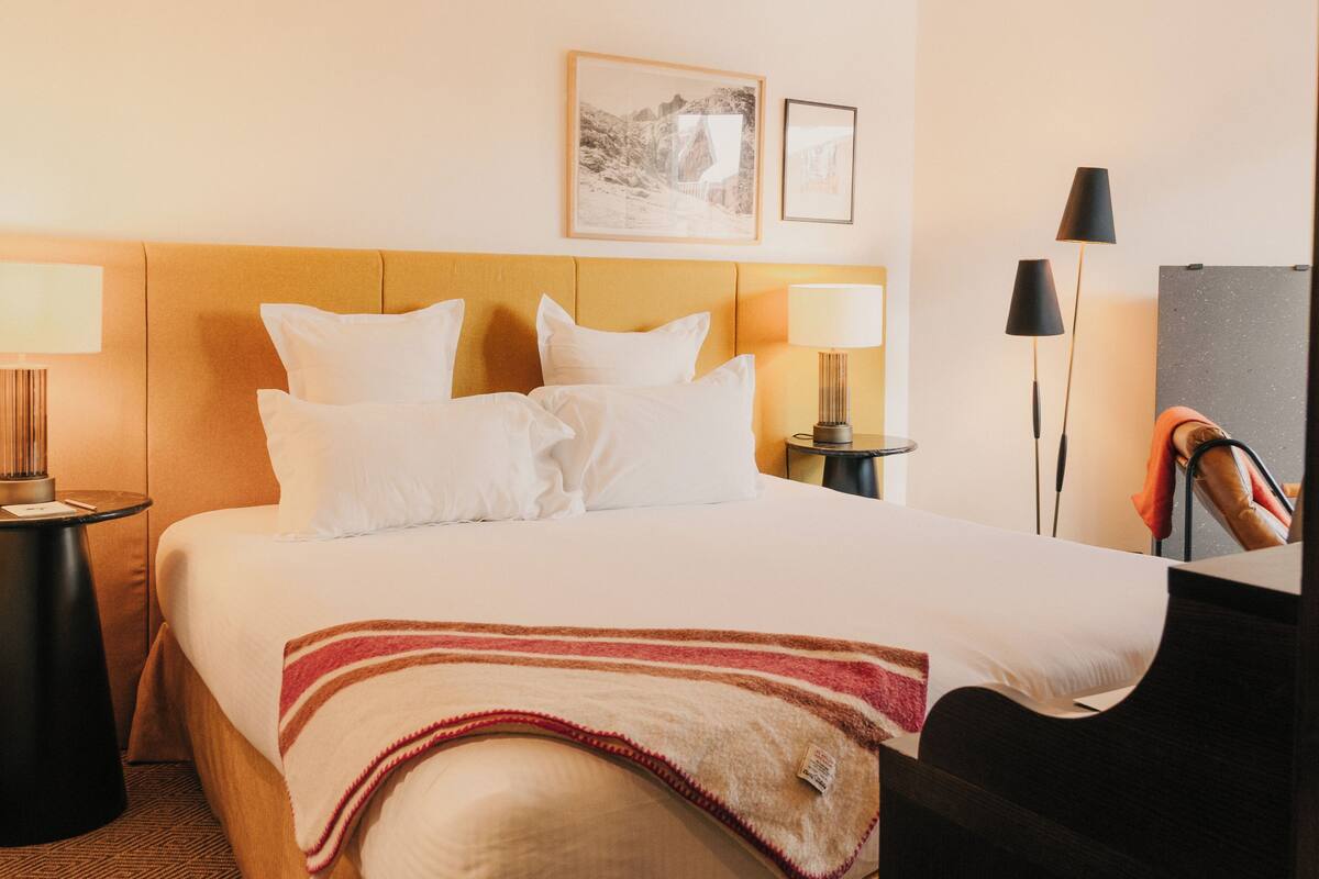 A hotel room with a bed, two bedside tables, a lamp, a chair, and two framed pictures on the wall.