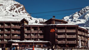 Een meerlaags houten gebouw met balkons, een bord met de tekst 'Le Val Thorens' en een besneeuwd berglandschap op de achtergrond.