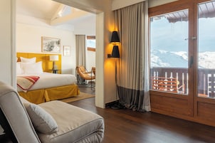 Habitación de hotel con cama, sofá, escritorio, lámpara, ventana con cortinas y vistas a una montaña nevada.