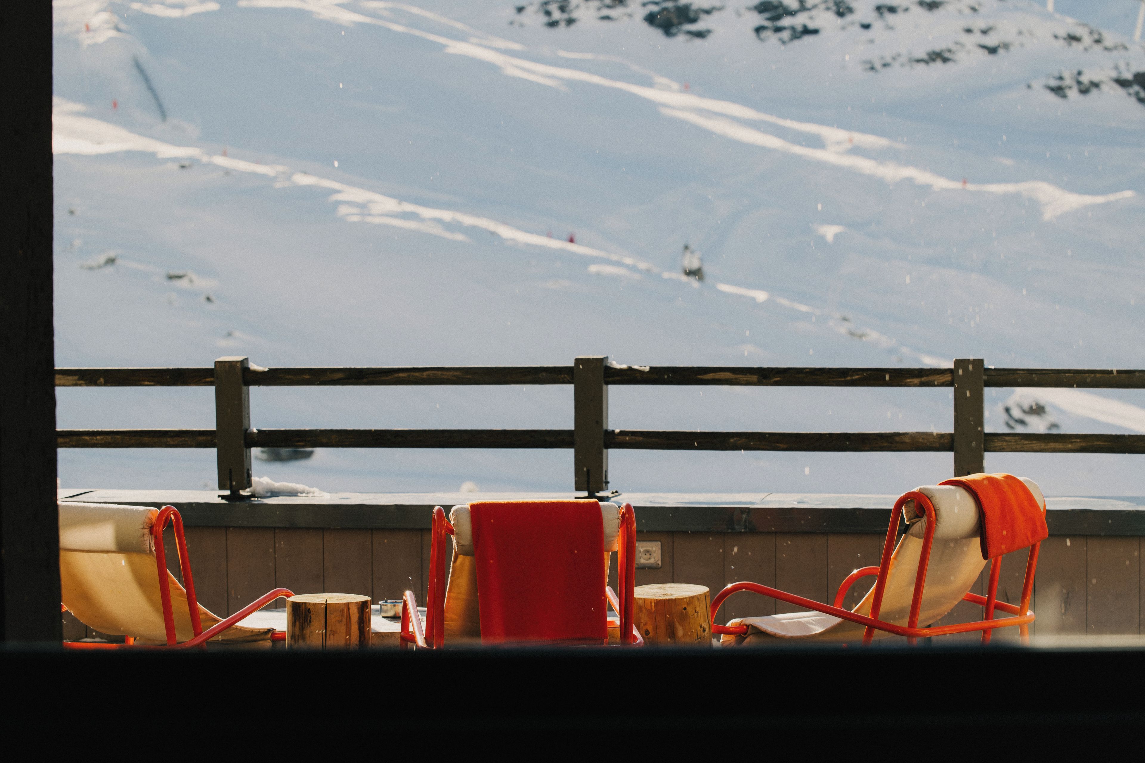 A view of a snowy landscape through a window with a wooden railing and two chairs.