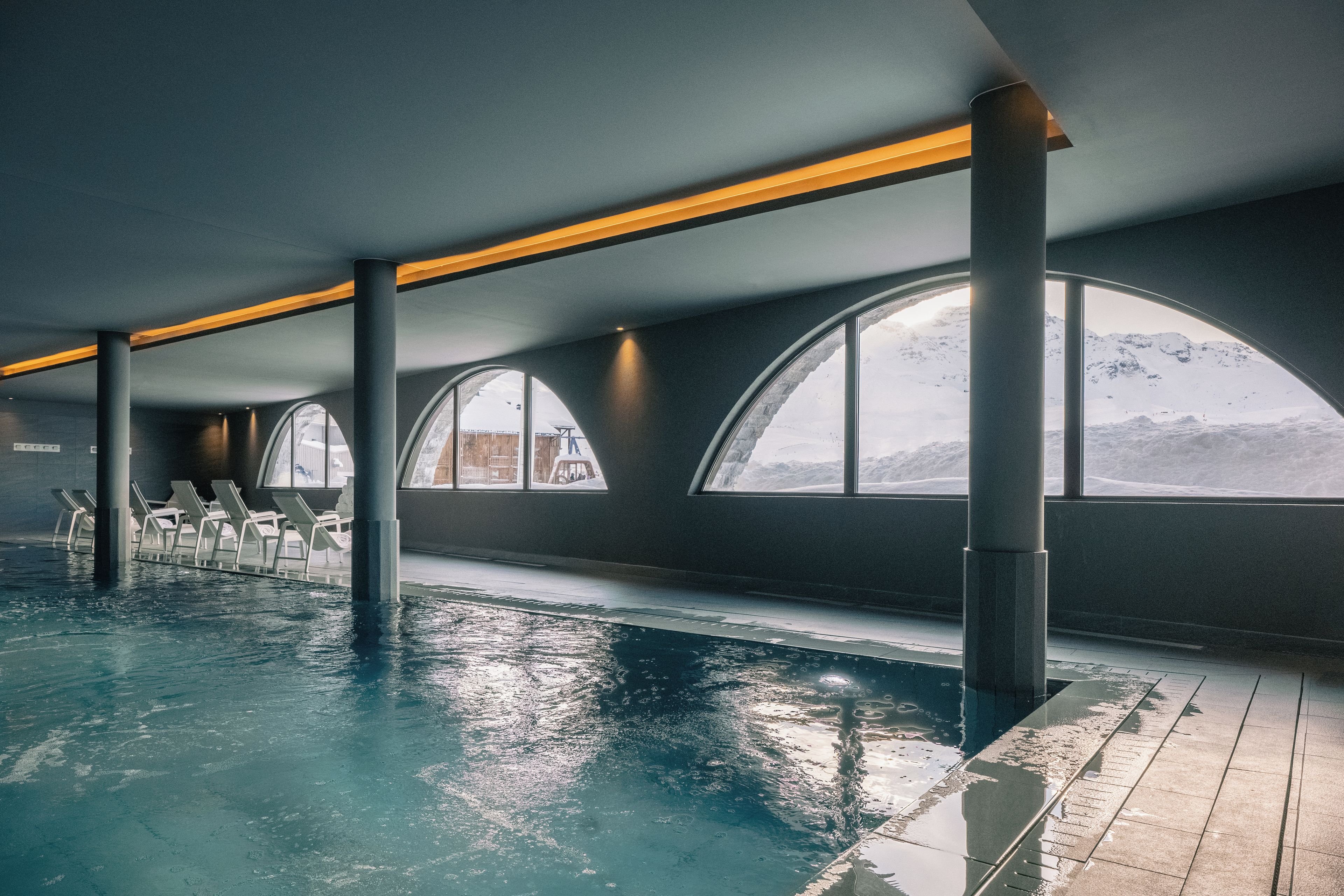 Una piscina interior con vistas a montañas a través de grandes ventanales, sillas de descanso y un diseño arquitectónico moderno.