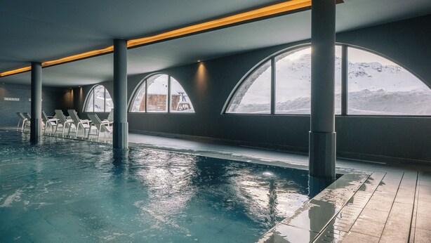 An indoor swimming pool with a view of mountains through large windows, lounge chairs, and a modern architectural design. - Le Val Thorens (Les Belleville)
