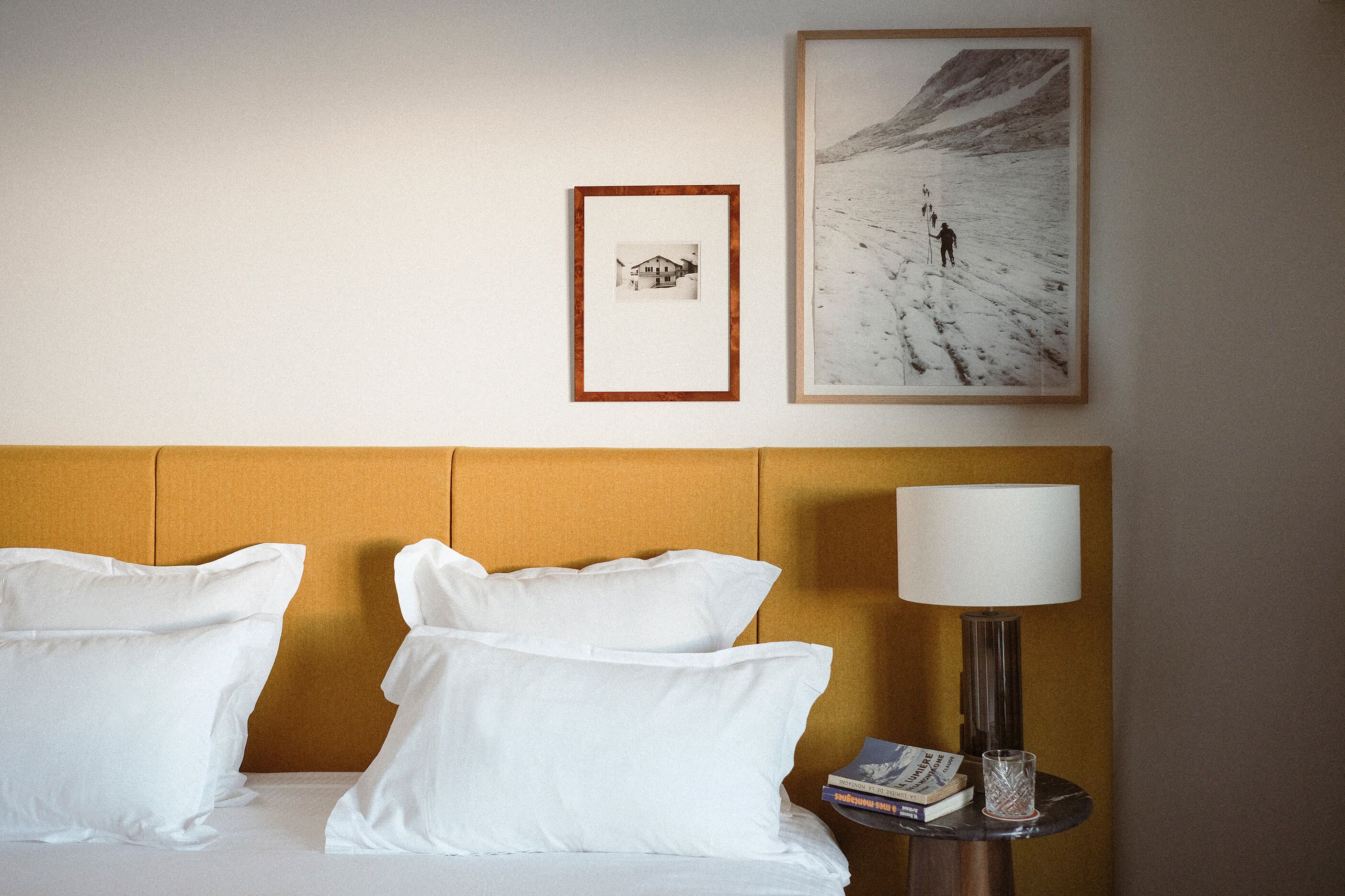 Habitación de hotel con cama, almohadas, mesita de noche con lámpara y dos cuadros en la pared.
