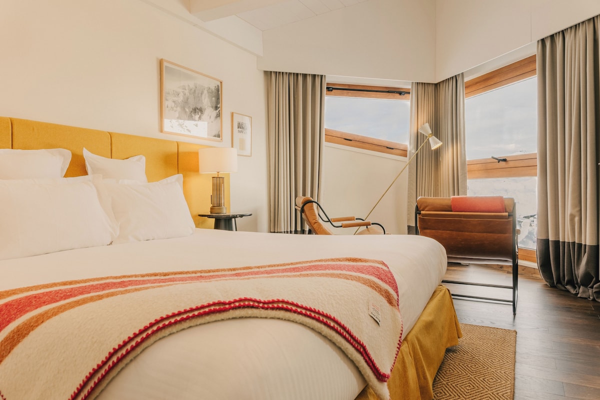 Habitación de hotel con cama, silla, escritorio, lámpara y ventana con cortinas.